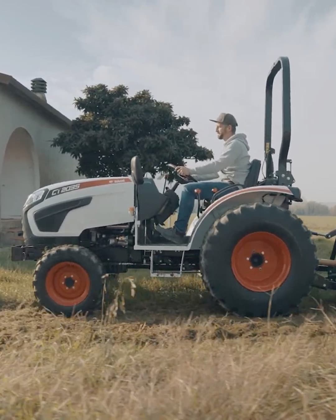 A person drives a Bobcat CT2035 Compact Tractor with a rear mower attachment across a grassy field near a building, demonstrating its capability for property and pasture maintenance.