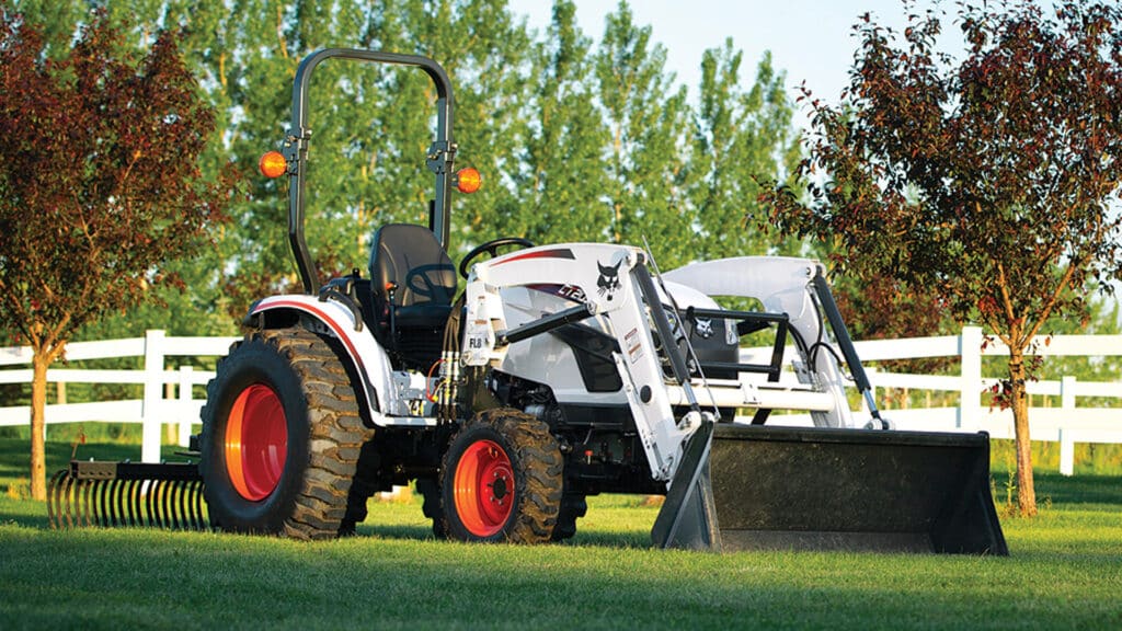 A Bobcat CT2035 Compact Tractor with a front-end loader and rear rake attachment parked on a well-maintained grassy property, surrounded by trees and a white fence.