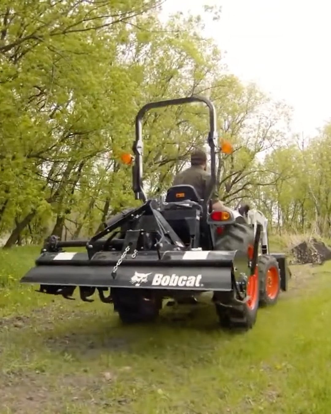 A person drives a Bobcat CT2040 Compact Tractor equipped with a rear rotary tiller attachment through a grassy, wooded area, ideal for soil preparation and rural land management.