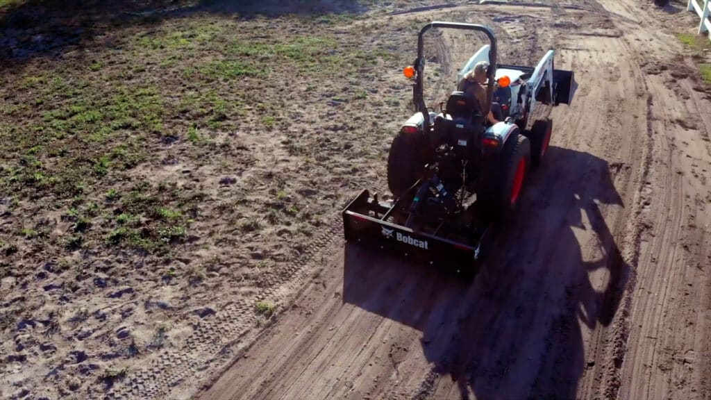 A person operates a Bobcat CT2040 Compact Tractor with a front-end loader and rear box blade, grading a dirt path on a rural property, demonstrating its versatility and power.