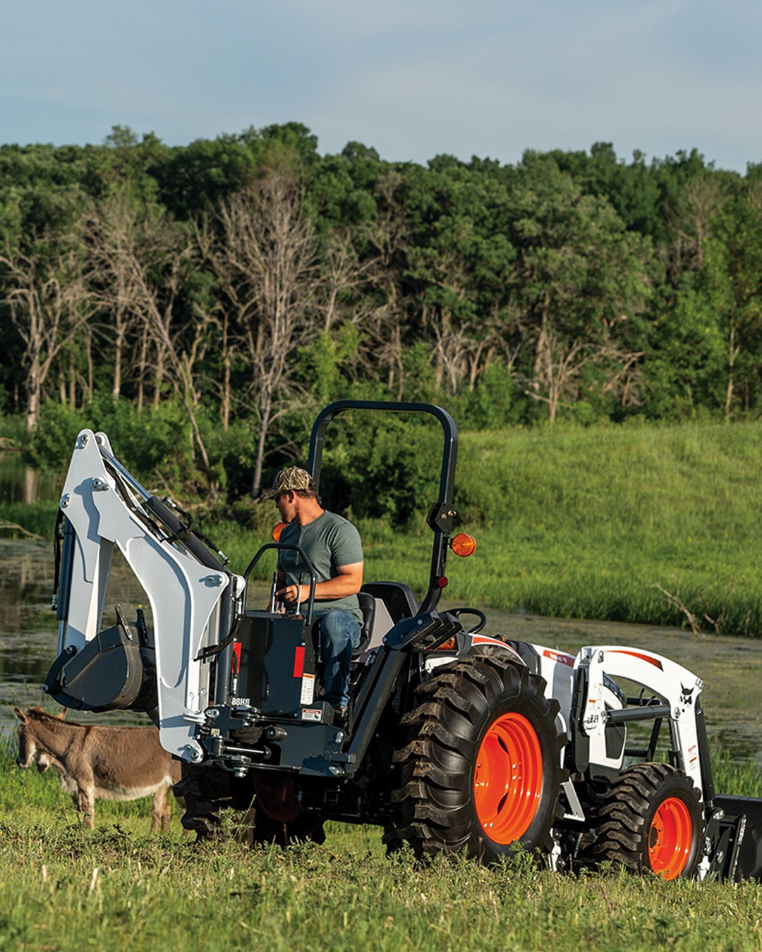 A person operates a Bobcat CT4045 Compact Tractor equipped with a backhoe and front-end loader, working near a pond with a donkey grazing nearby, ideal for farm and property maintenance.