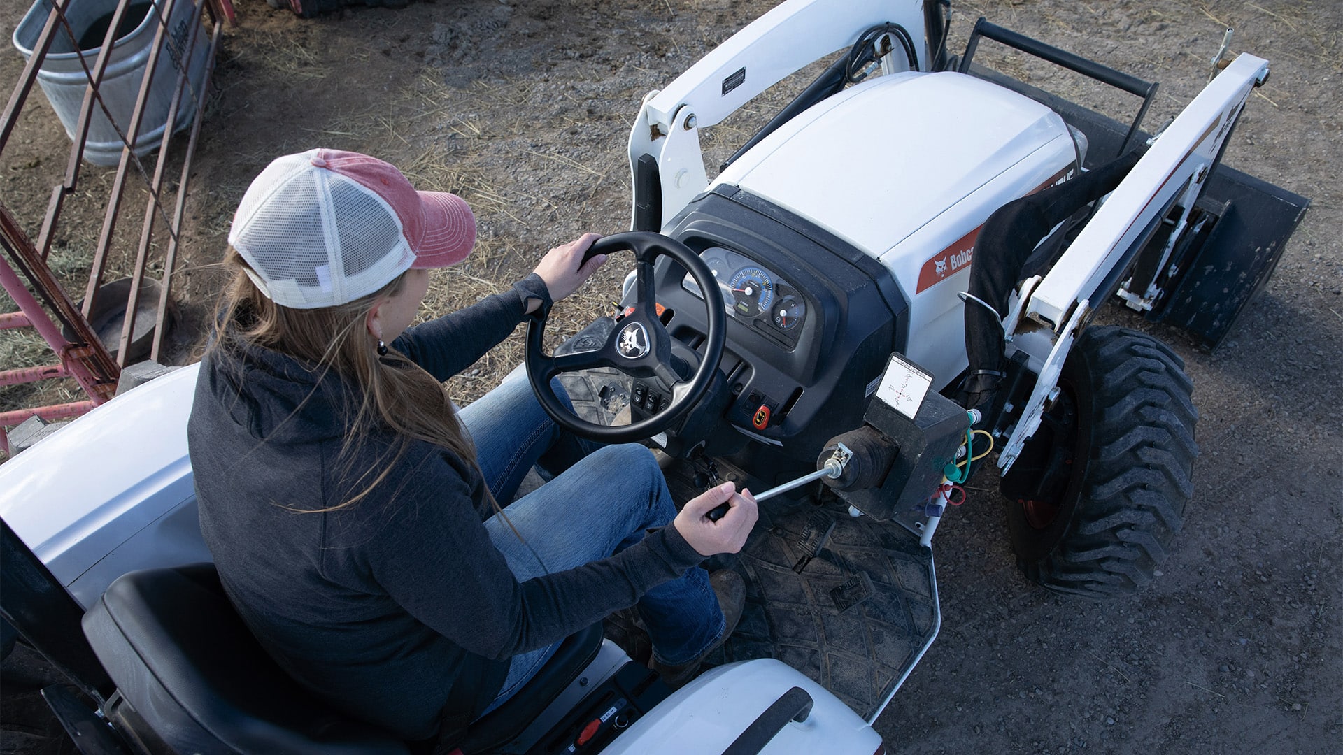 A person operates a Bobcat CT4045 Compact Tractor, viewed from above, showing the control panel, steering wheel, and loader joystick in action on a dirt-covered worksite.