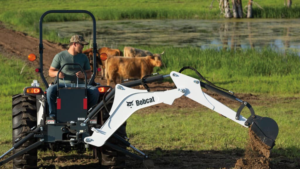 A person operates a Bobcat CT4045 Compact Tractor with a BH86 backhoe attachment, digging soil near a pond while cattle graze in the background, ideal for farm and land management work.