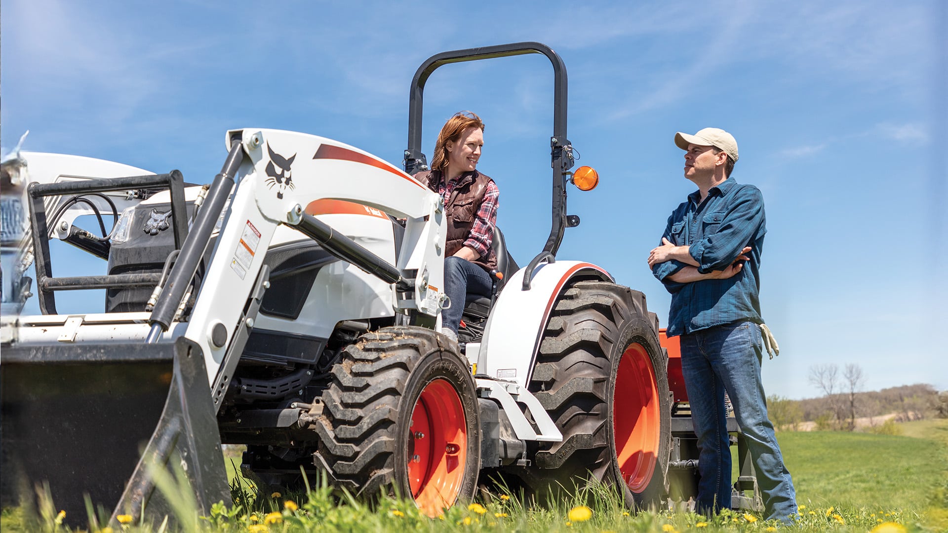 A person sits on a Bobcat CT4055 Compact Tractor with a front-end loader, talking to another person standing nearby in a grassy field under a clear blue sky, showcasing the tractor in a rural work environment.