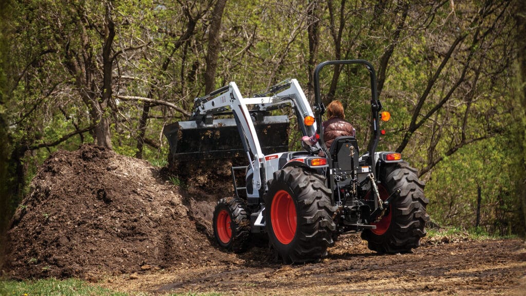A person operates a Bobcat CT4055 Compact Tractor with a front-end loader, moving a large pile of soil in a wooded area, demonstrating its strength and efficiency for demanding rural tasks.