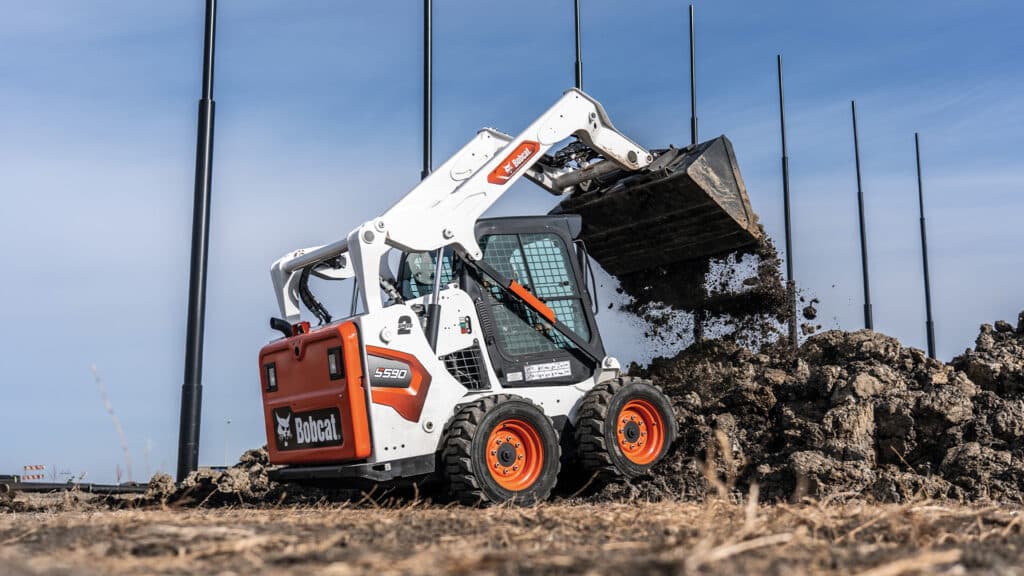 Bobcat S590 Skid Steer Loader lifting and dumping soil on a construction site under a clear blue sky.