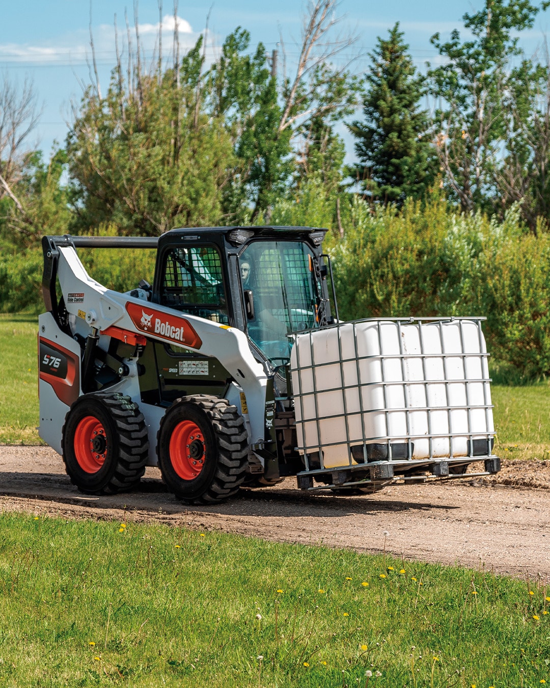 Bobcat S76 Skid Steer Loader transporting a large water tank on a pallet, demonstrating its material handling capability on a gravel path surrounded by lush greenery.