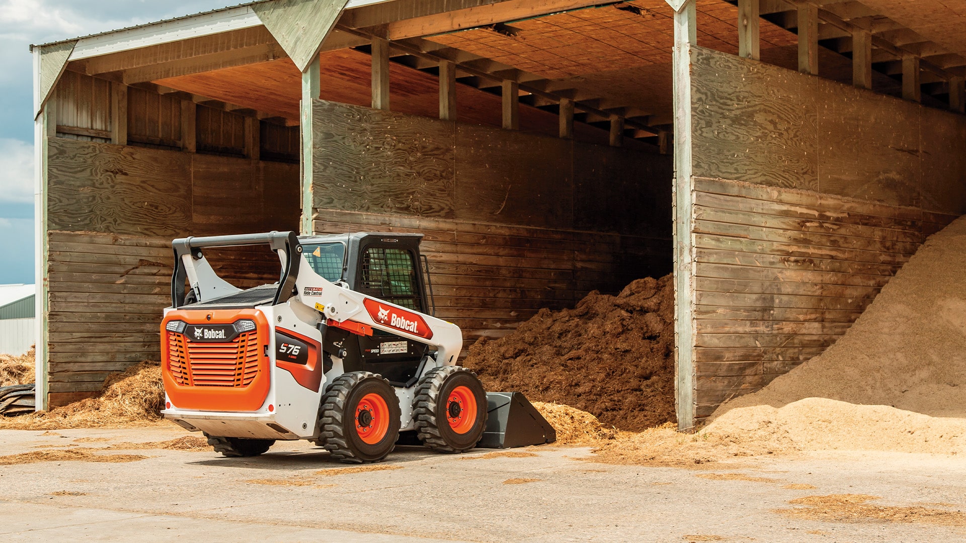 Bobcat S76 Skid Steer Loader parked near a large pile of straw in a storage shed, ready for efficient material handling and barn cleanup.