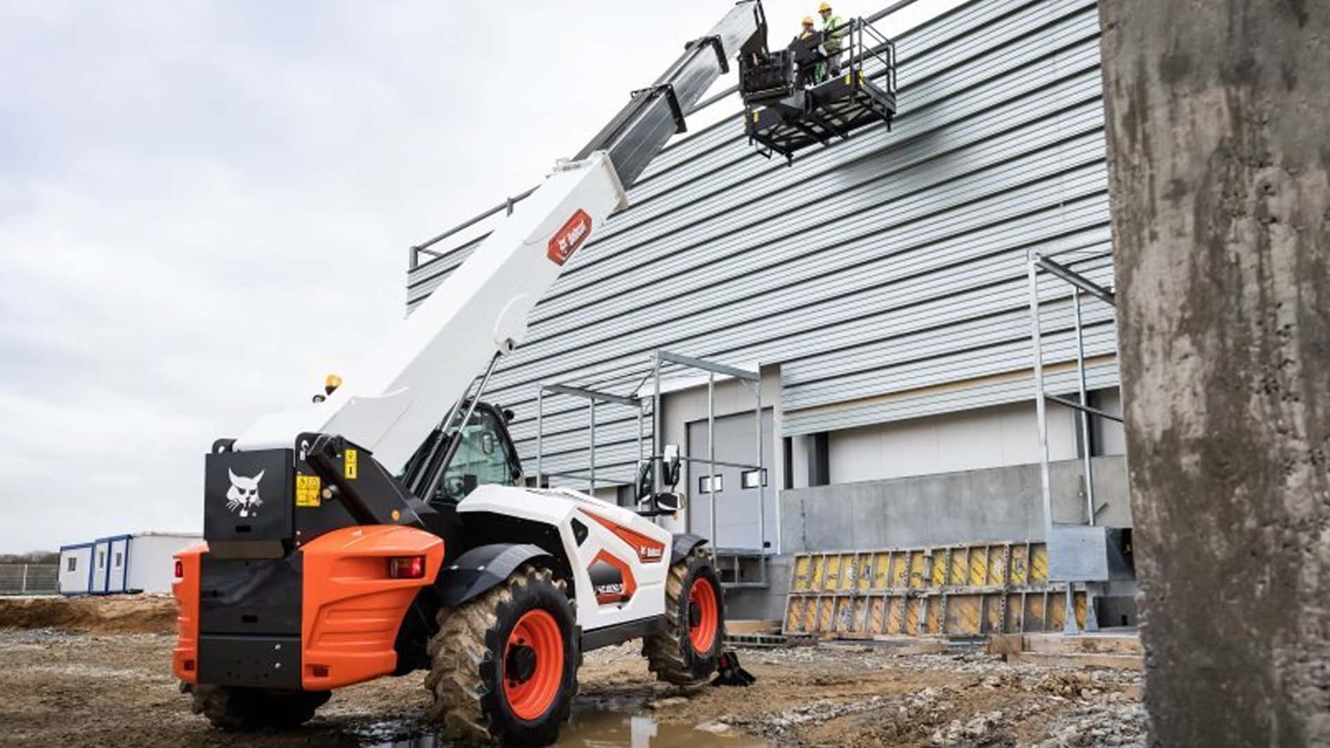A Bobcat T40.180 Construction Telehandler lifting two workers in a safety platform to access the side of a large industrial building, demonstrating its precision, stability, and versatility for high-reach tasks on construction sites.