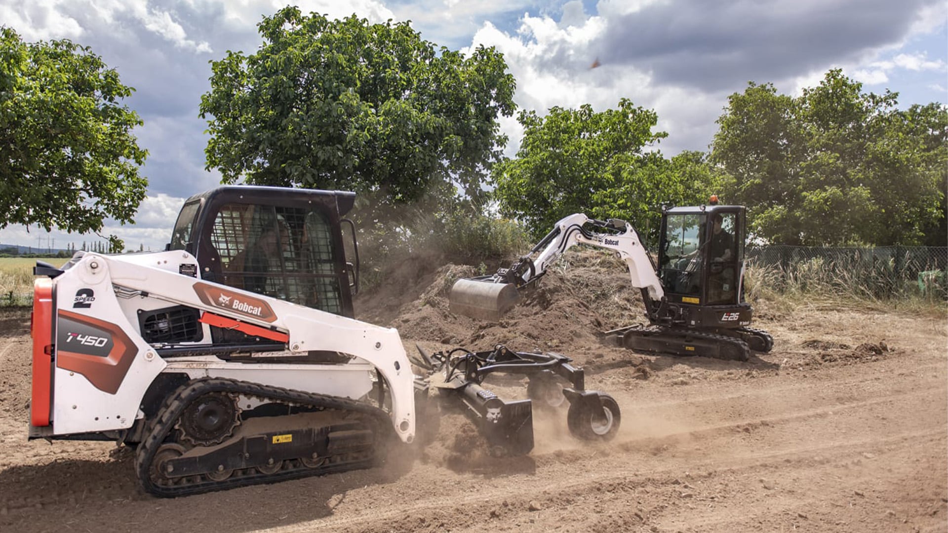 Bobcat T450 Track Loader with a soil conditioner attachment working alongside a Bobcat E26 Compact Excavator on a dusty construction site surrounded by greenery and cloudy skies.