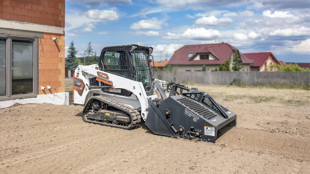 Bobcat T450 Track Loader with a landscape rake attachment levelling soil in a residential yard, surrounded by newly constructed houses and a clear, partly cloudy sky.