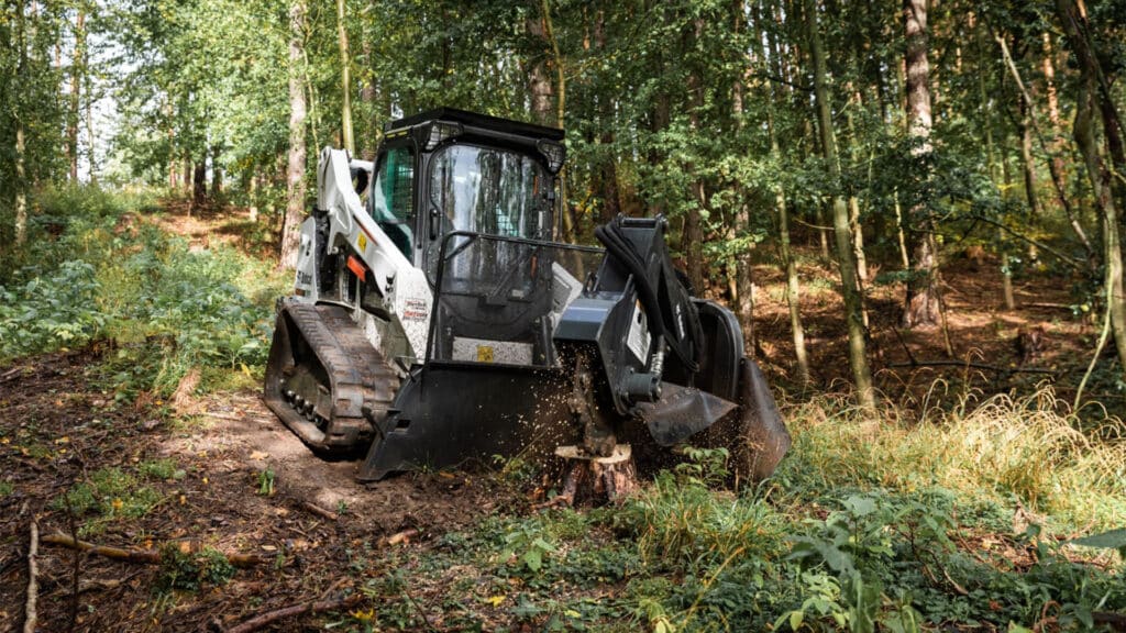 Bobcat T590 Track Loader clearing a forest trail with a stump grinder attachment, surrounded by dense woodland and lush green foliage.