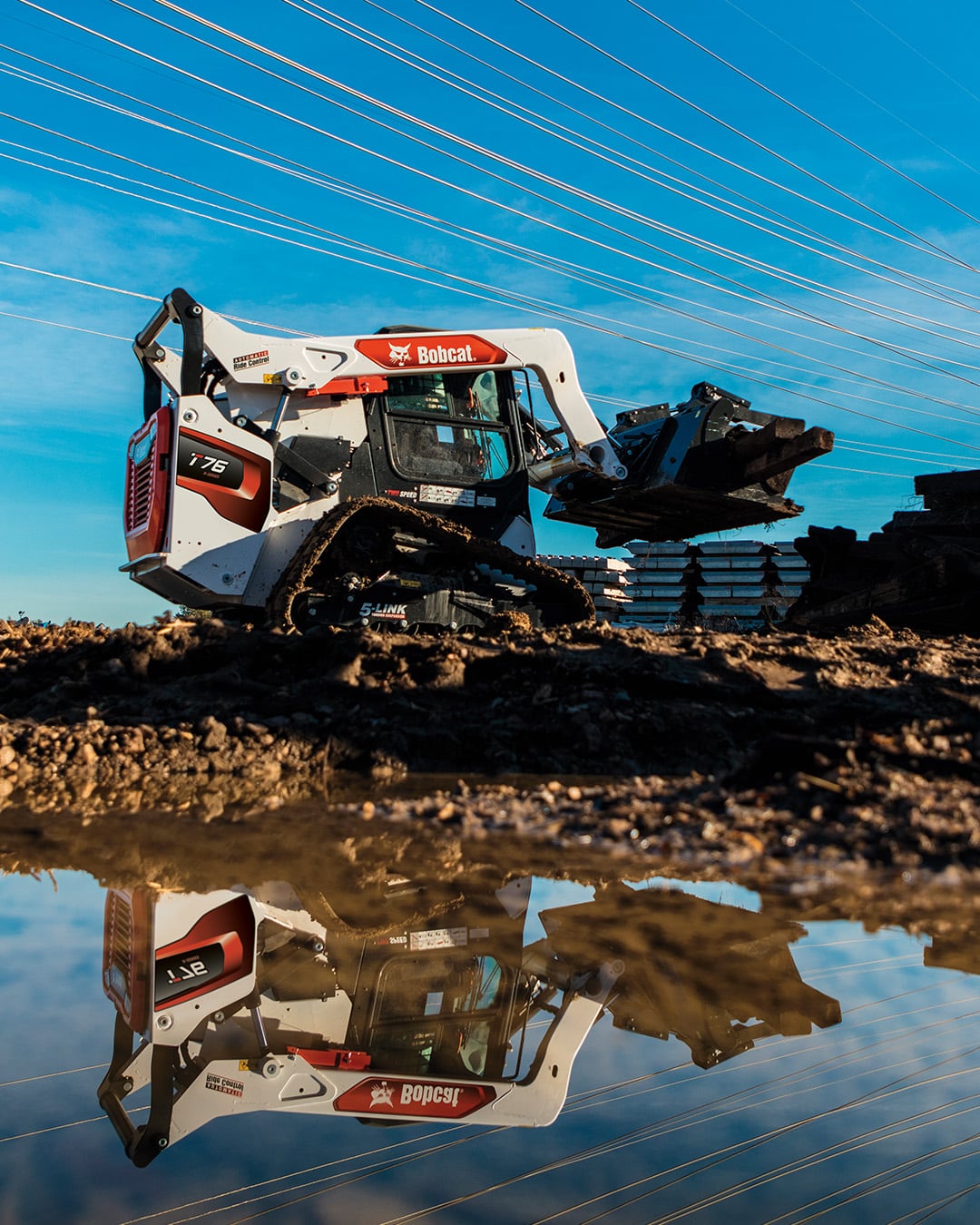 A Bobcat T76 Track Loader lifting materials on a rugged worksite with power lines overhead, reflected in a puddle for a striking visual effect.
