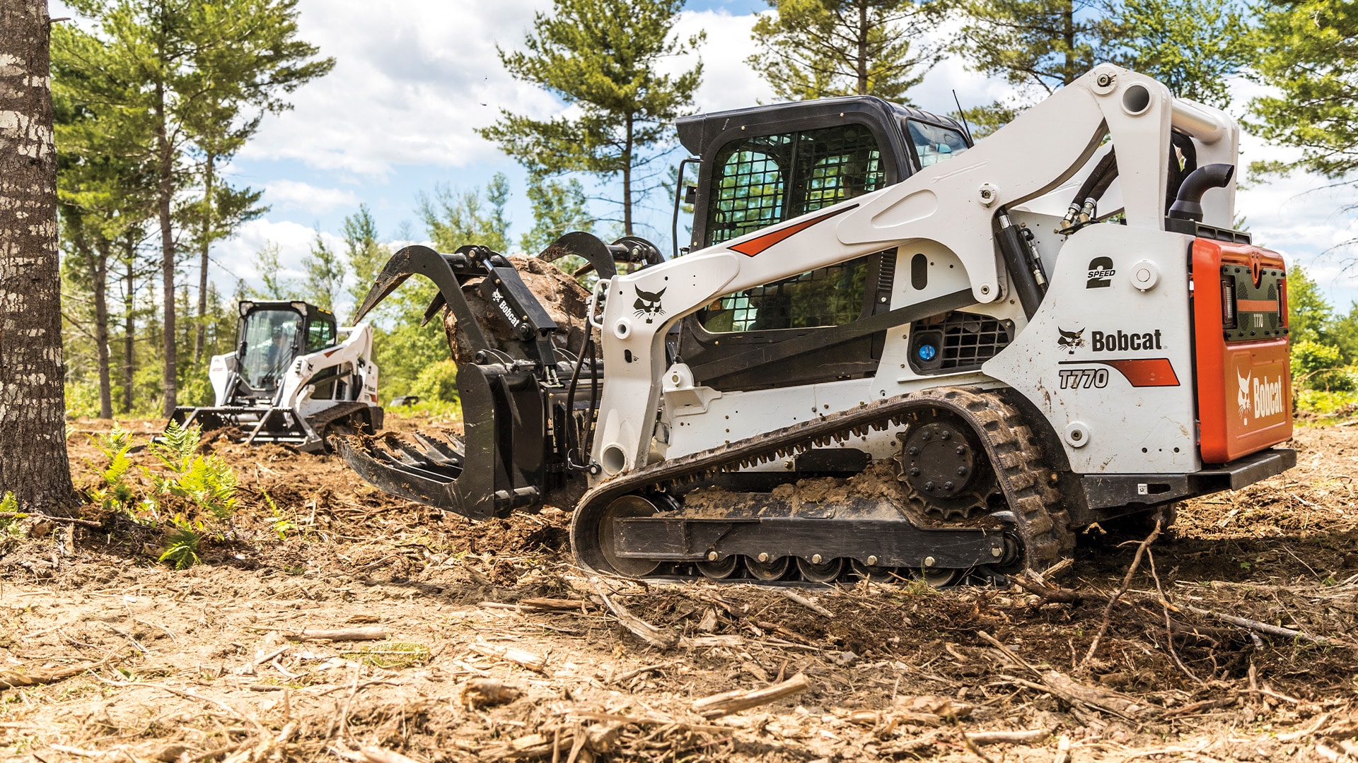 A Bobcat T770 Track Loader equipped with a grapple attachment, clearing brush and debris in a forested area, with another Bobcat loader in the background working on site preparation.