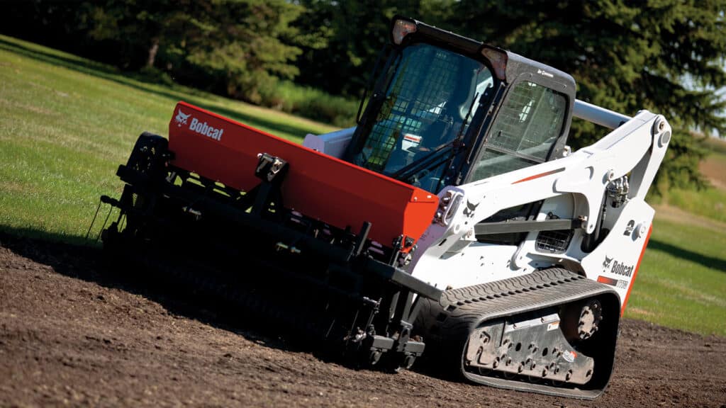 A Bobcat T770 Track Loader equipped with a seeder attachment, preparing soil for landscaping and turf installation on a grassy field.