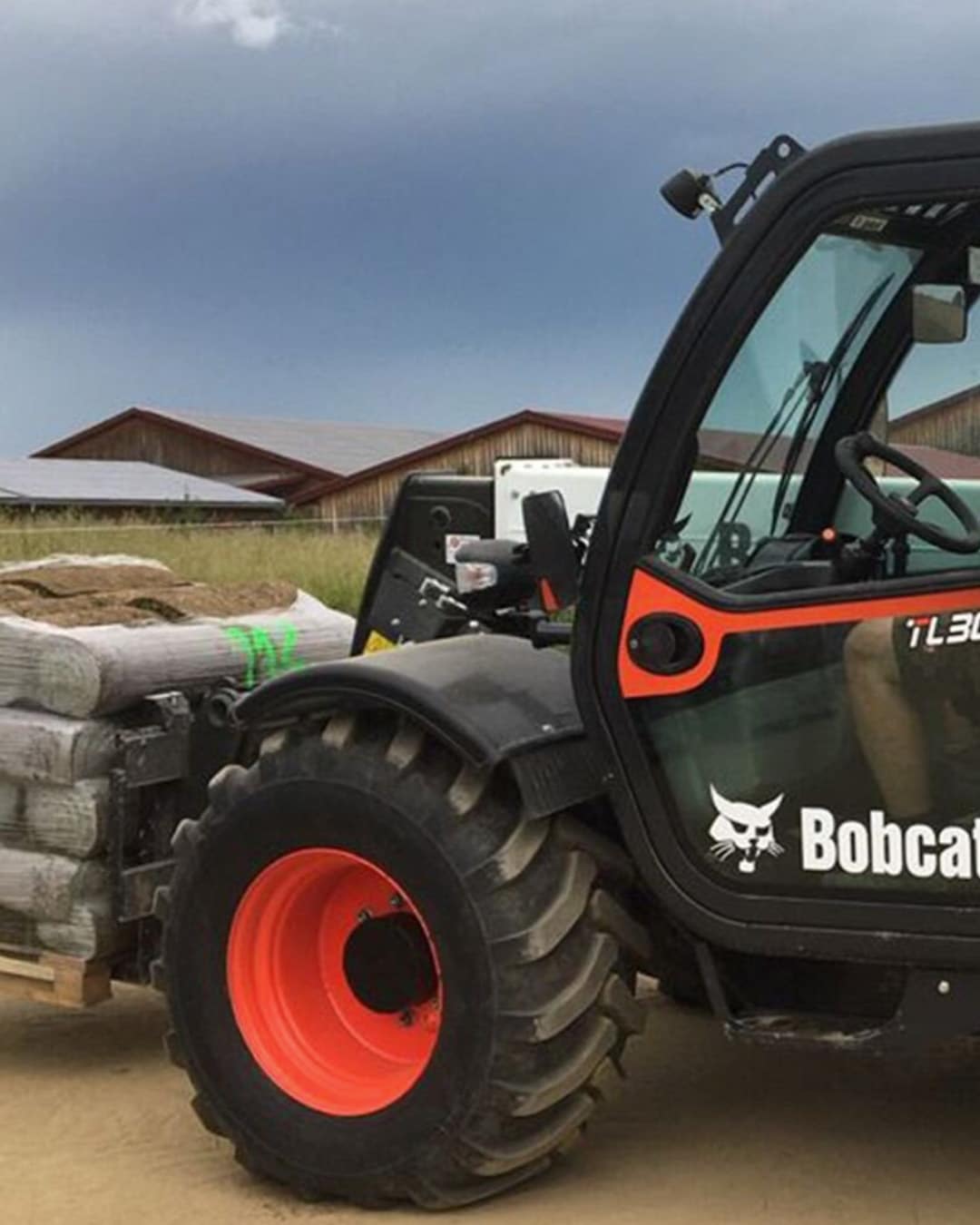 A Bobcat TL30.60 Agricultural Telehandler transporting a pallet of feed or supplies in a rural setting, with barns and open farmland in the background.