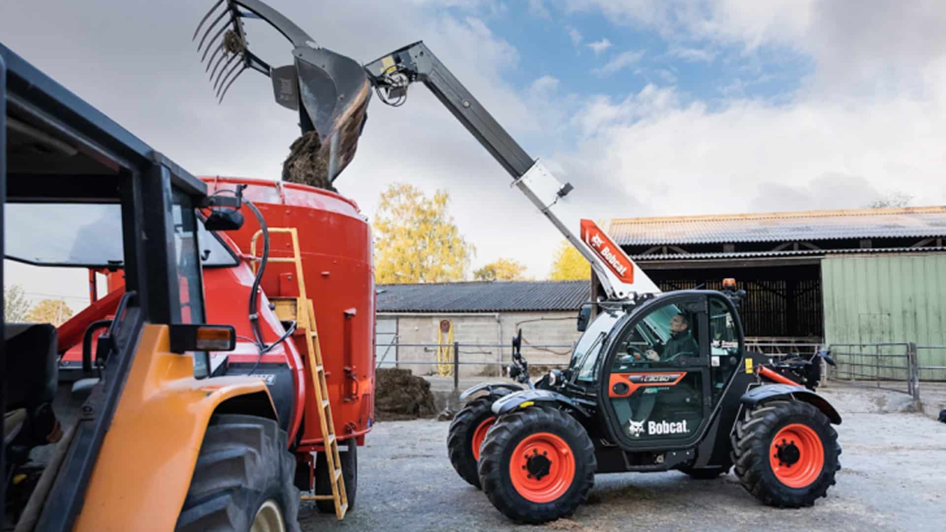 A Bobcat TL30.60 Agricultural Telehandler loading hay or silage into a red feed mixer wagon on a working farm, with a tractor and farm sheds in the background.