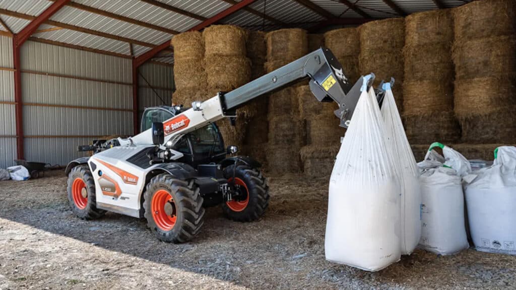 A Bobcat TL30.60 Agricultural Telehandler lifting large bulk feed bags inside a hay shed, surrounded by stacked round hay bales and farm supplies.