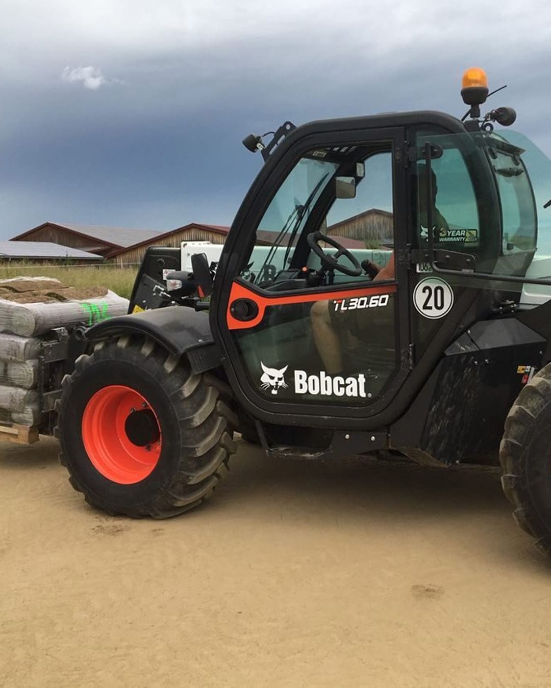 A Bobcat TL30.60 Construction Telehandler transporting a pallet of construction materials on a dirt worksite, with sheds and a cloudy sky in the background.