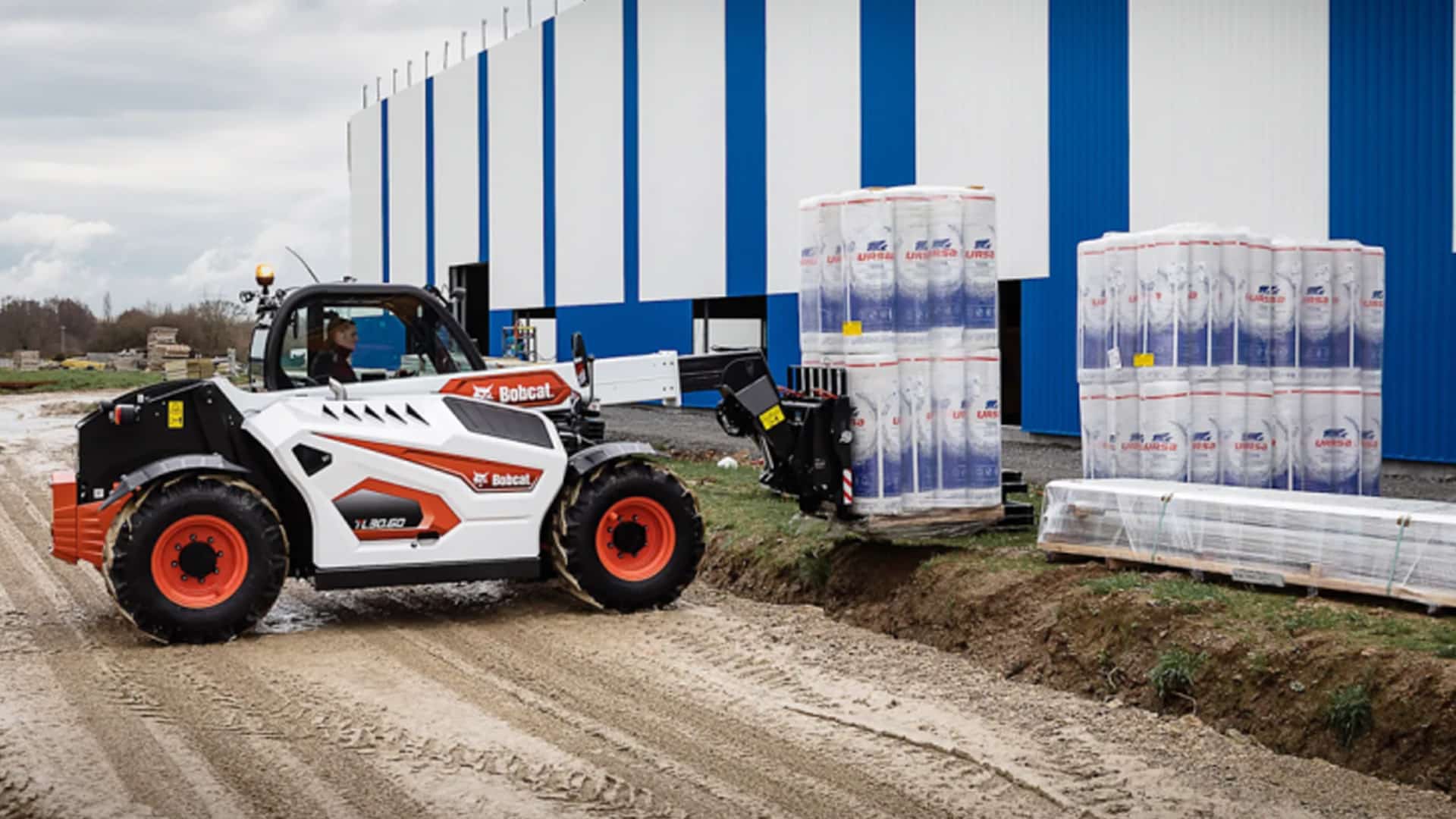A Bobcat TL30.60 Construction Telehandler operating on a commercial construction site, lifting a pallet of insulation material with its pallet fork attachment near a modern industrial building.