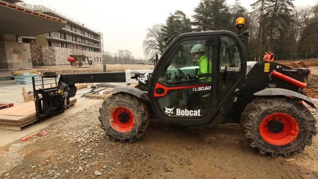 A Bobcat TL30.60 Construction Telehandler in use on an active building site, operated by a worker in safety gear, lifting construction materials with a pallet fork attachment.