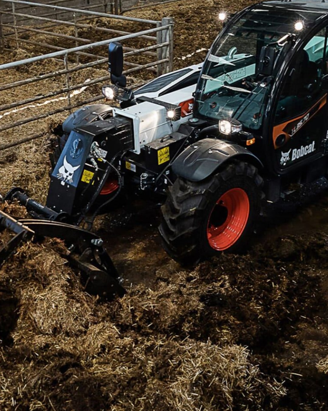 A Bobcat TL35.70 Agricultural Telehandler clearing straw and manure inside a livestock pen, equipped with a grapple bucket attachment.