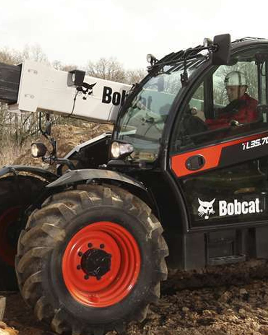 A Bobcat TL35.70 Construction Telehandler operating on a muddy job site, with a worker inside the cab wearing a hard hat, ready to lift or move materials using the extended boom arm.