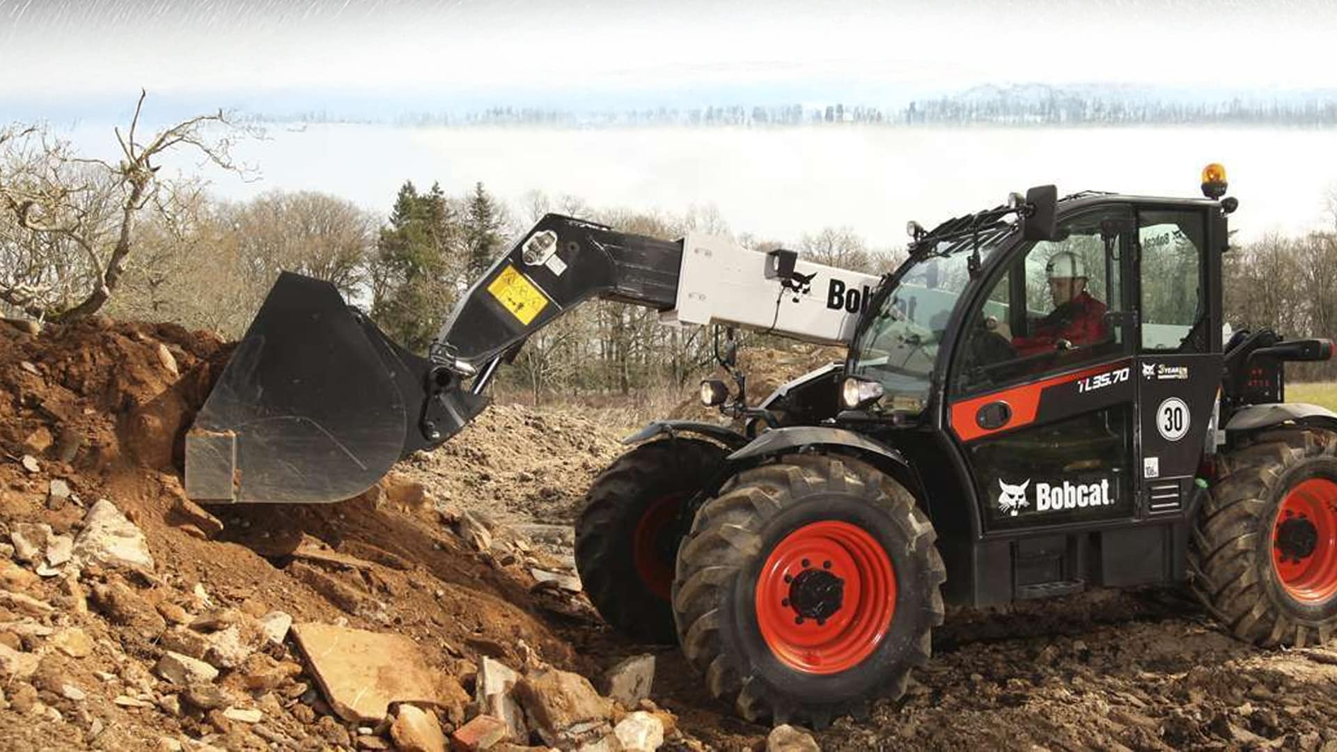 A Bobcat TL35.70 Construction Telehandler scooping up loose soil and rocks on a rugged construction site, operated by a worker wearing a hard hat inside the cab.