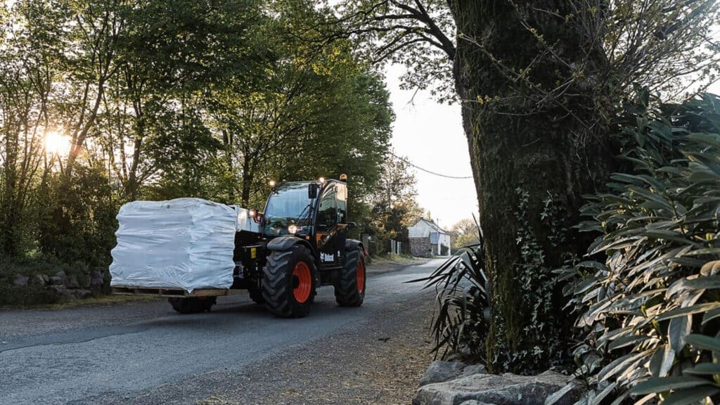 A Bobcat TL35.70 Construction Telehandler transporting a pallet of wrapped materials along a rural road at sunrise, surrounded by trees and greenery.