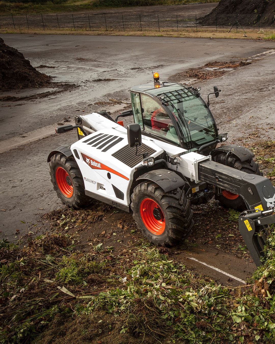A Bobcat TL38.70HF Agricultural Telehandler with a grab attachment, clearing green waste and vegetation on a muddy outdoor site.