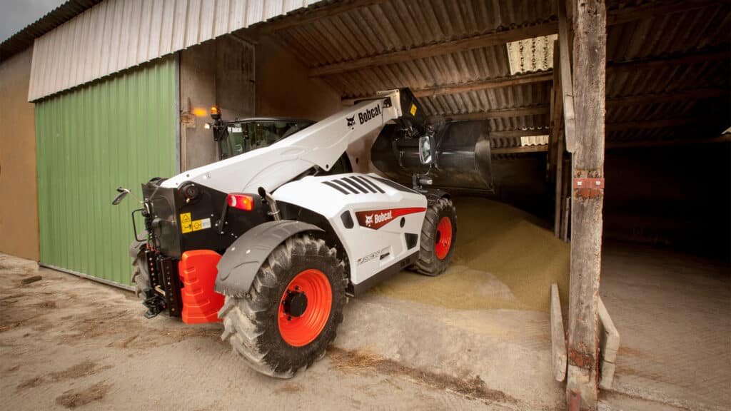 A Bobcat TL38.70HF Agricultural Telehandler working inside a farm shed, using a bucket attachment to move a large pile of grain or feed.