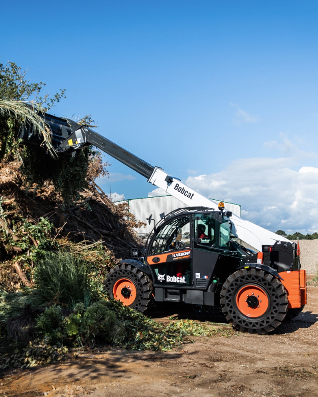 A Bobcat TL43.80HF Agricultural Telehandler in action, clearing a large pile of green waste using a grapple bucket on a sunny day at a composting or agricultural facility, with blue skies and industrial buildings in the background.