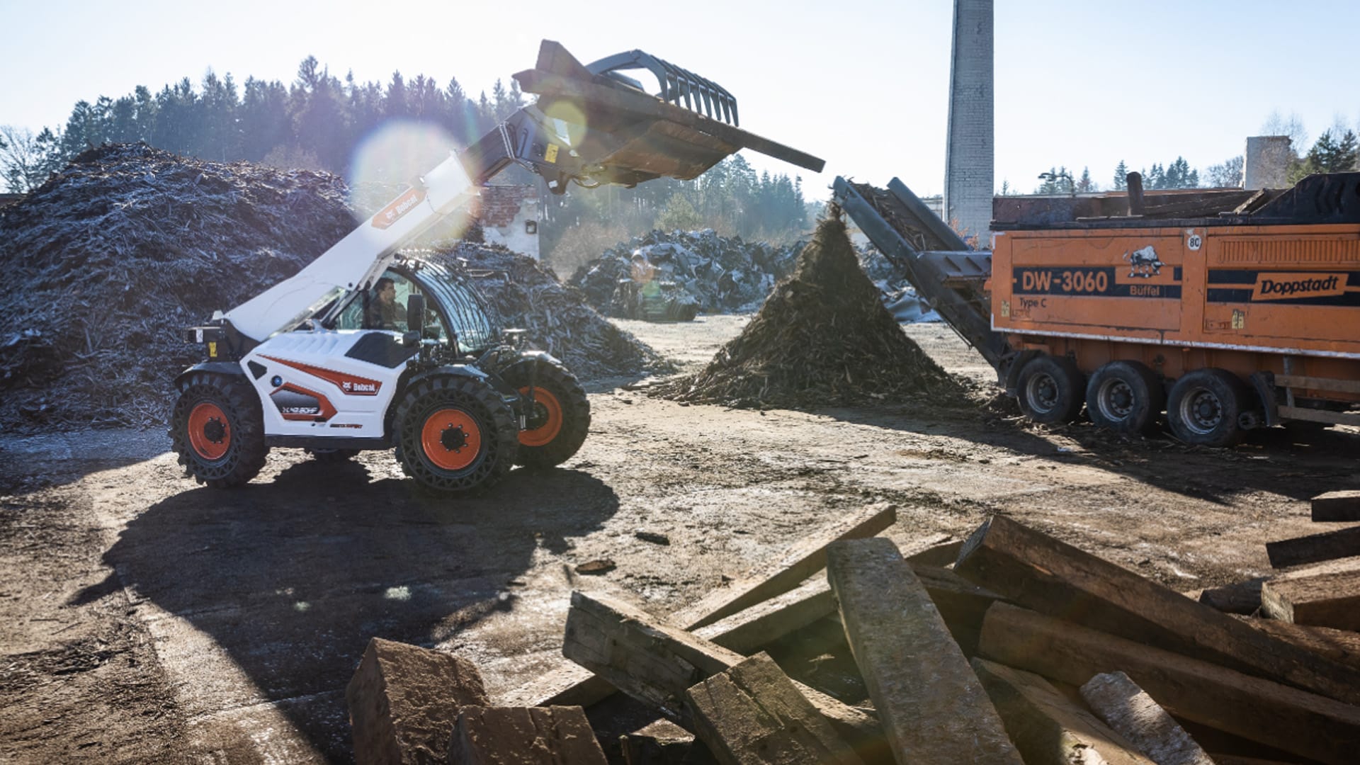 A Bobcat TL43.80HF Agricultural Telehandler operating on a recycling or composting site, lifting debris with a grapple bucket attachment near a large orange industrial machine, surrounded by piles of organic waste and wood under bright daylight.