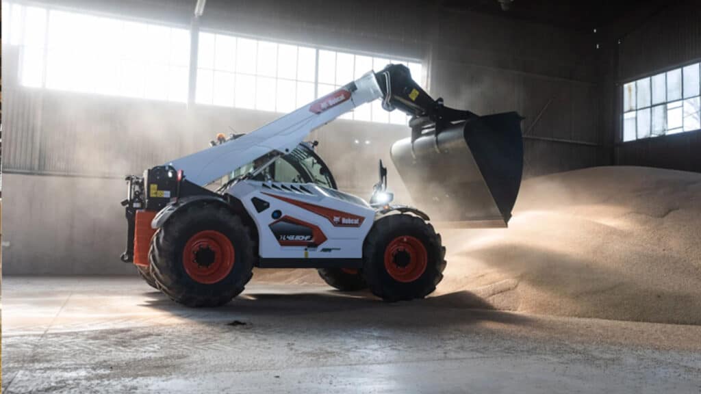 A Bobcat TL43.80HF Agricultural Telehandler working inside a storage shed, using a bucket to manage a large pile of grain or feed, with sunlight streaming through the windows and dust suspended in the air.