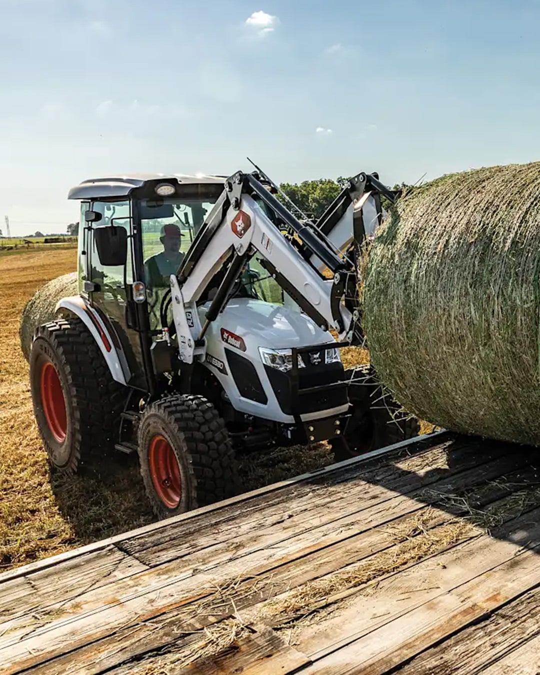 Bobcat UT6570 Utility Tractor loading a large round hay bale onto a wooden trailer bed in a dry paddock on a sunny day.