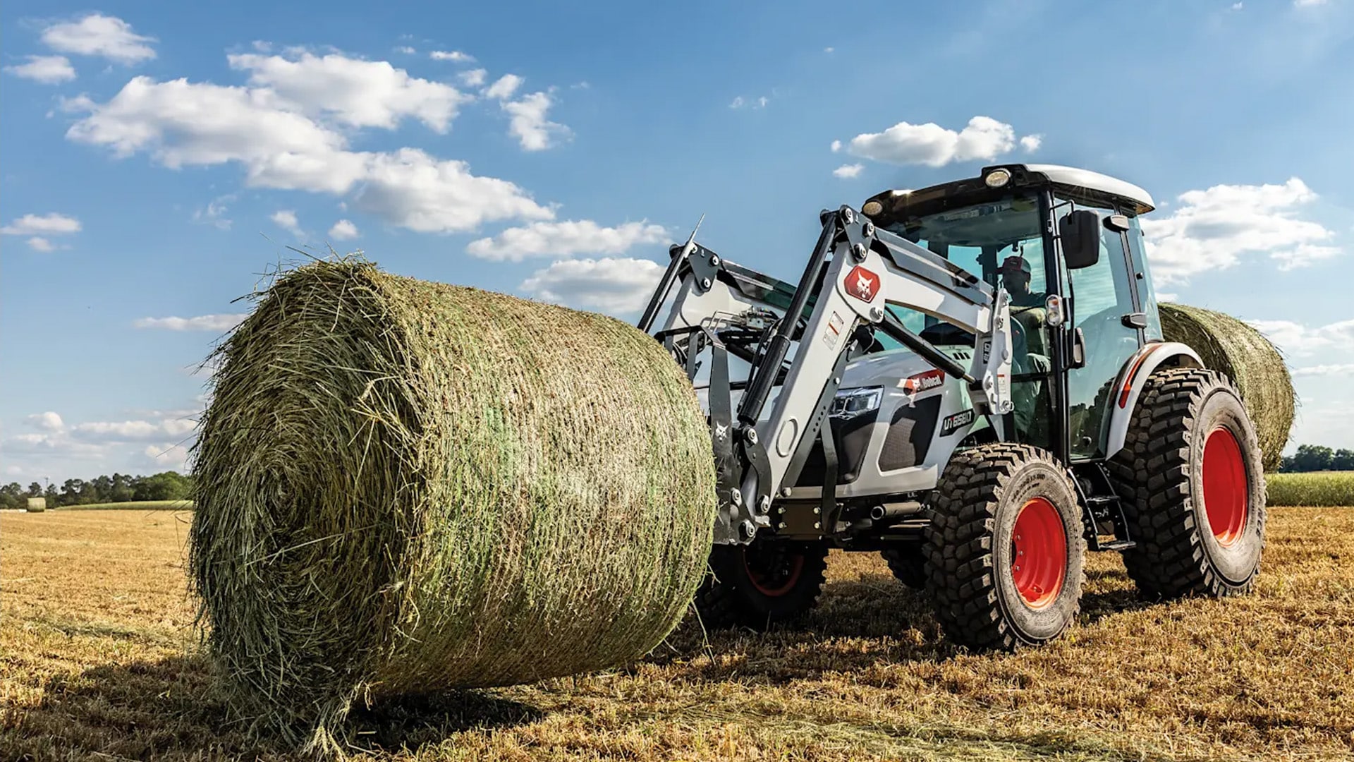 Bobcat UT6570 Utility Tractor with cab lifting a large round hay bale in a dry, harvested field under a blue sky with scattered clouds.