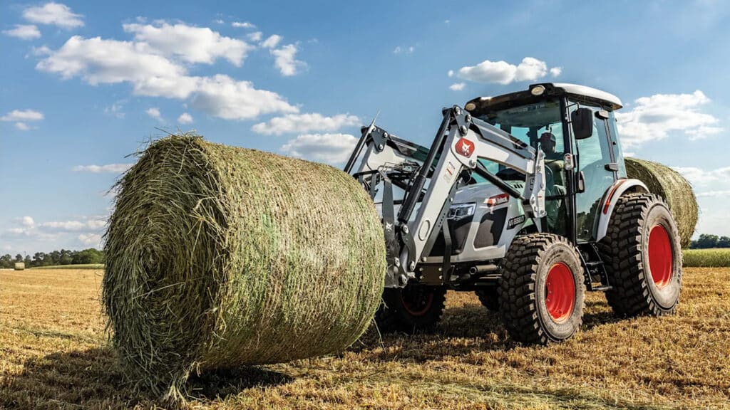 Bobcat UT6580 Utility Tractor equipped with a front-end loader, preparing to lift a large round hay bale in a harvested field under a blue sky.