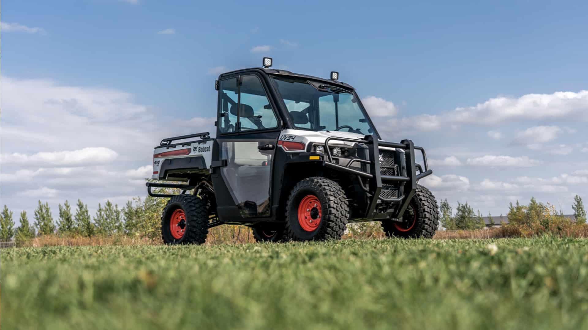 A Bobcat UV34 UTV with an enclosed cab and rugged tyres is parked on a grassy field under a blue sky, highlighting its durability and readiness for heavy-duty outdoor work.