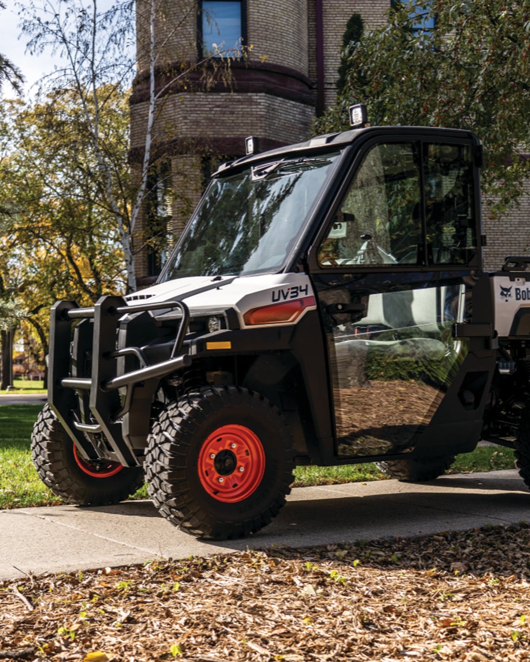 A fully enclosed Bobcat UV34 UTV with a protective front bumper and rugged tyres is parked on a sidewalk near a brick building, highlighting its utility for property maintenance and urban landscaping.