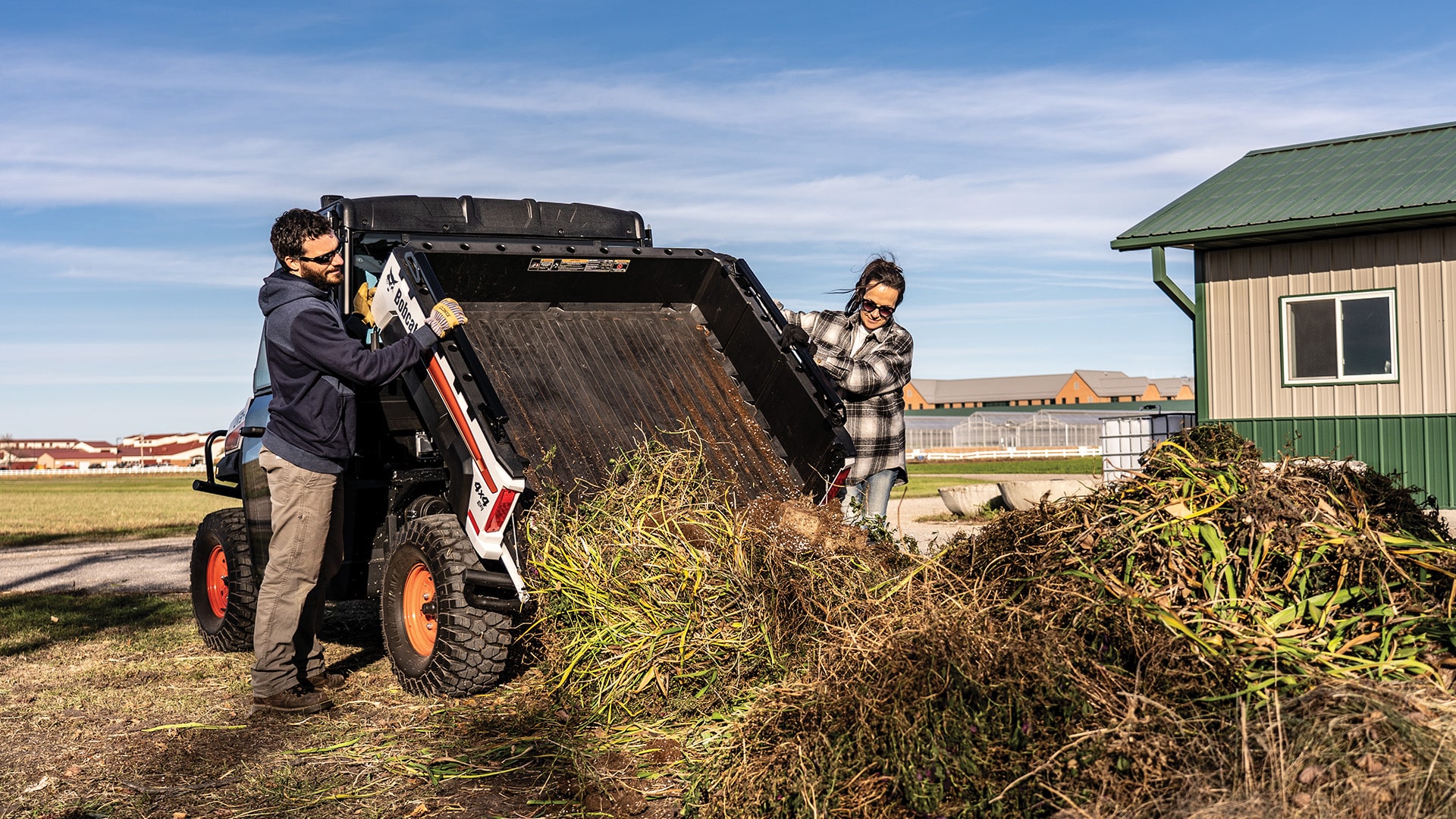 Two people unload a large pile of garden waste from the rear dump bed of a Bobcat UV34 UTV on a sunny property, demonstrating its practical use for landscaping and farm cleanup.