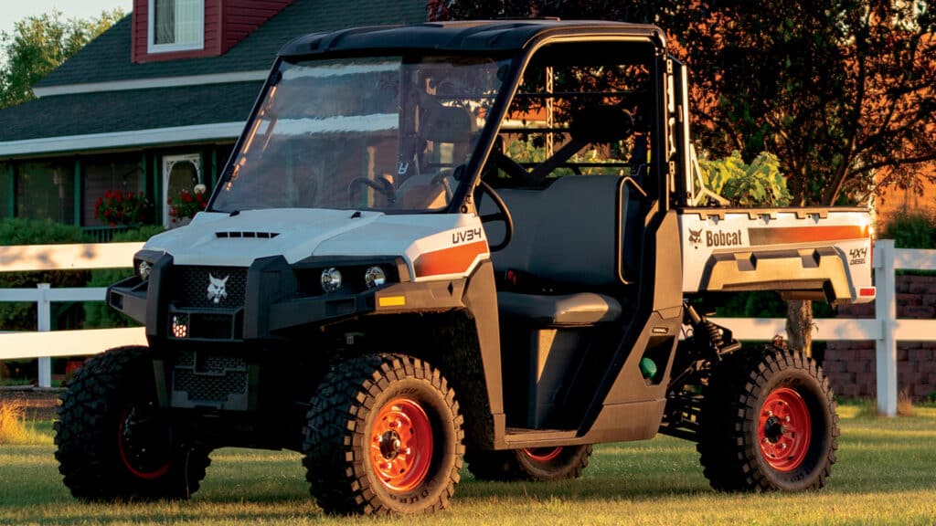 A Bobcat UV34 UTV with rugged tires and a white-and-black body is parked on a grassy lawn near a farmhouse, showcasing its 4x4 diesel power and utility for property maintenance.
