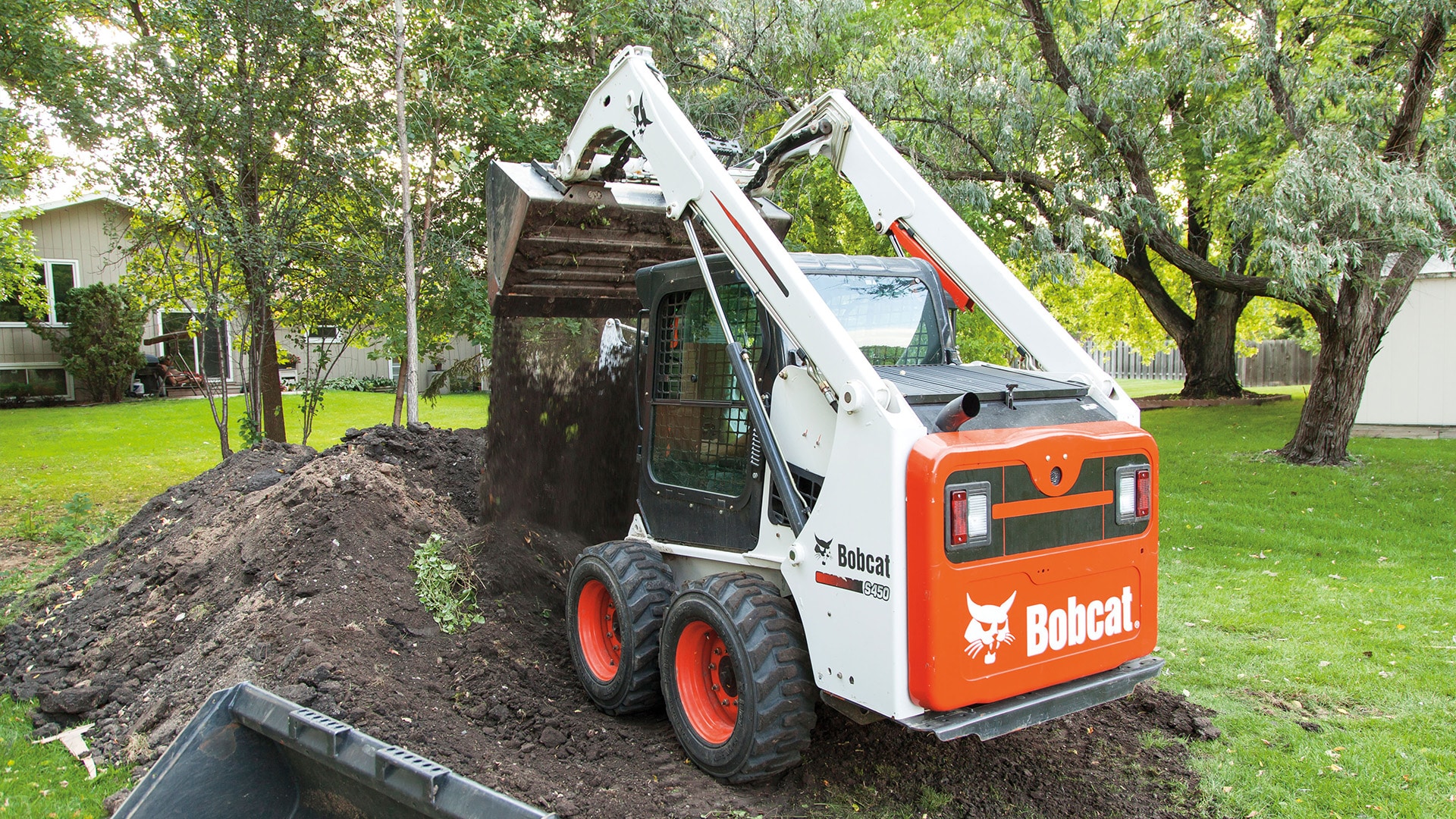 Bobcat S450 Skid Steer Loader moving a load of soil in a residential backyard, efficiently handling landscaping tasks with ease.