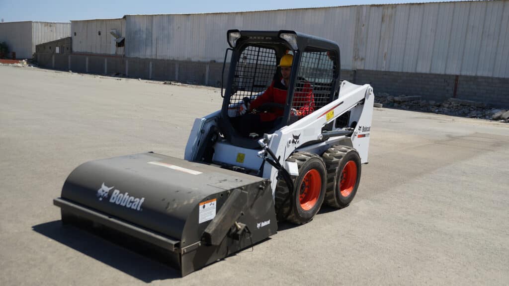 Bobcat S450 Skid Steer Loader equipped with a sweeper attachment, efficiently cleaning a large concrete surface on a construction site.