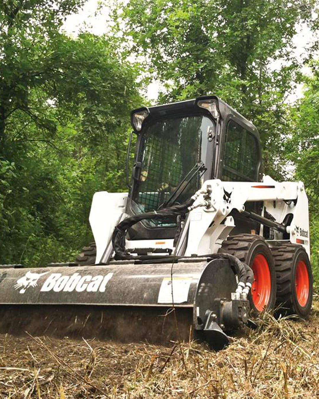 Bobcat S550 Skid Steer Loader with a forestry mulcher attachment, clearing dense vegetation in a wooded area.