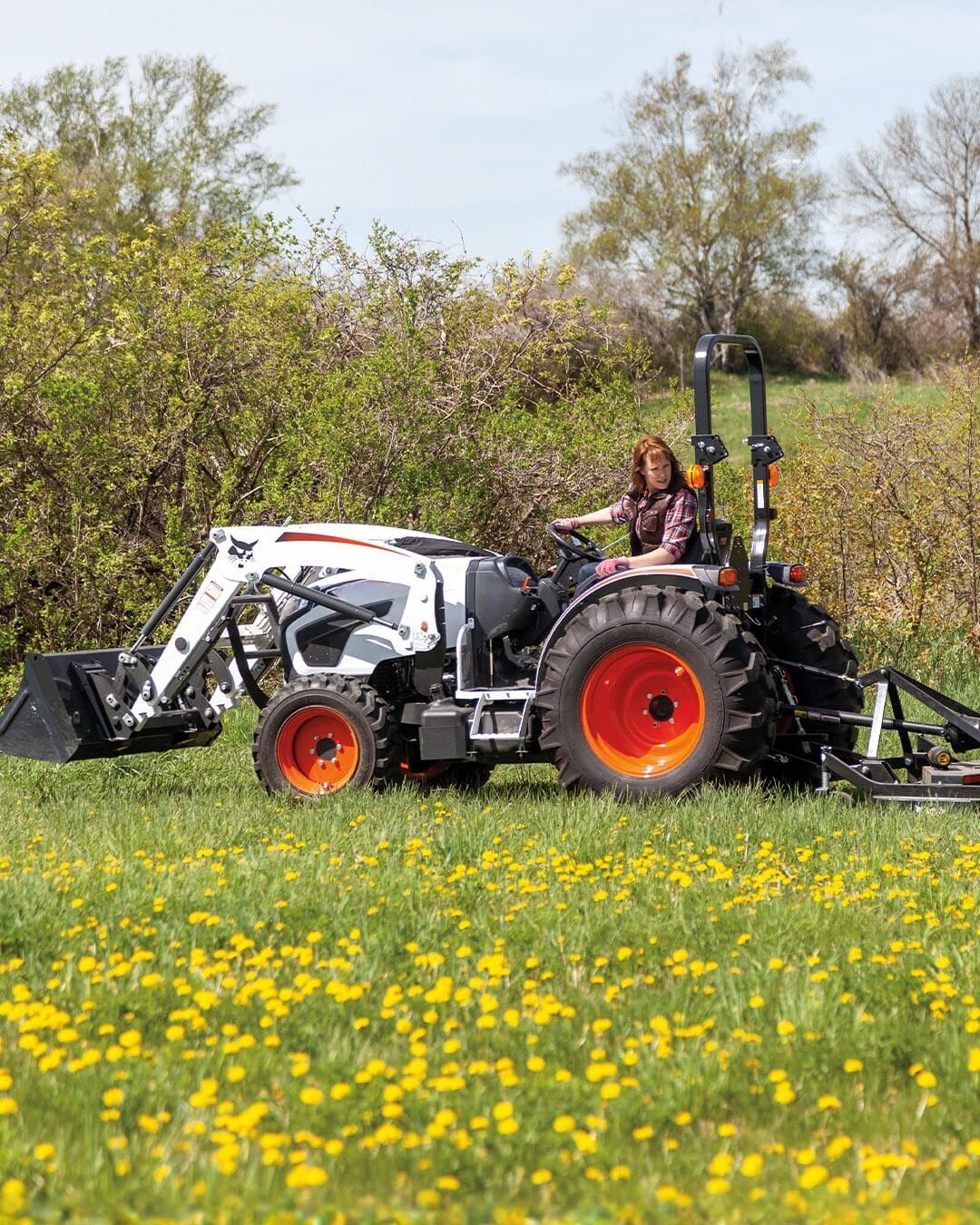 A person drives a Bobcat Small Tractor with a front-end loader and rear attachment through a field of yellow wildflowers, demonstrating its versatility for rural property maintenance.