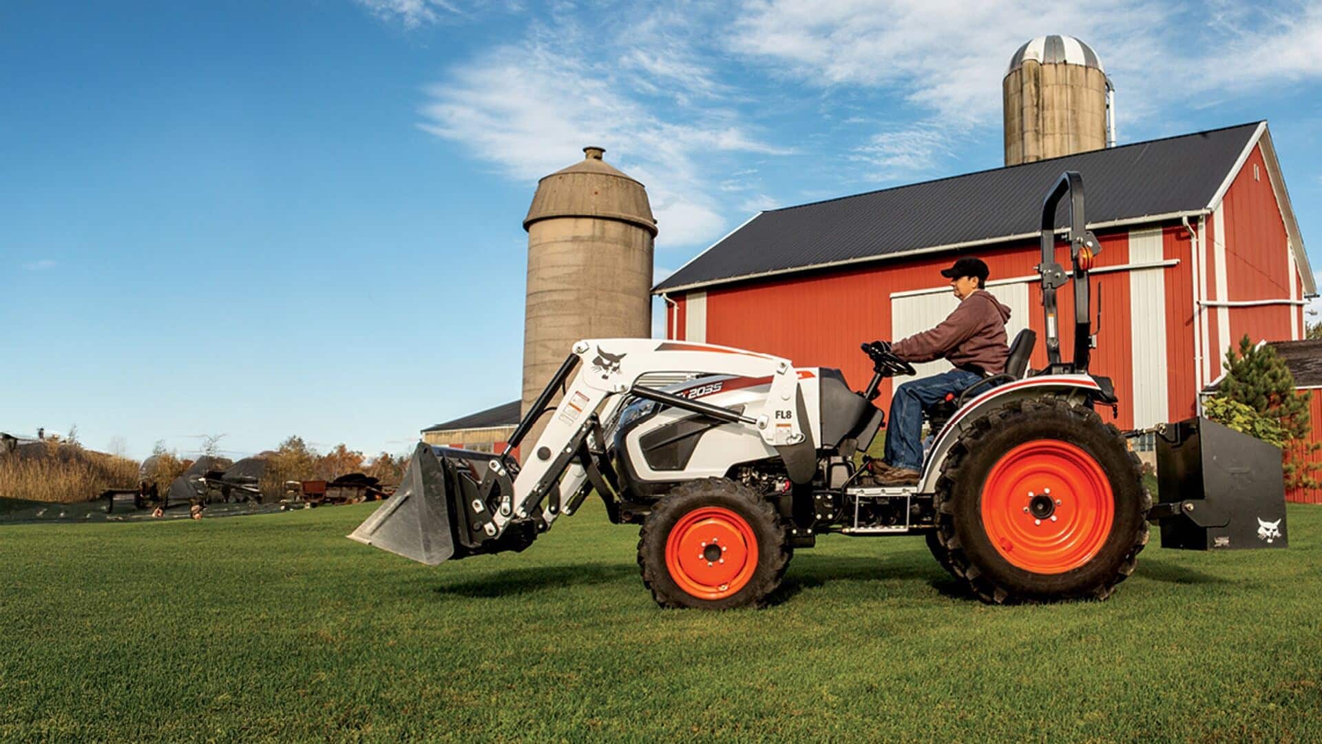 A person operates a Bobcat Small Tractor with a front-end loader on a grassy farm near a red barn and silos, highlighting its capability for light agricultural work.