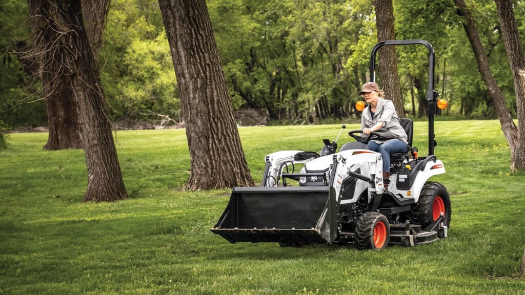A person drives a Bobcat Small Tractor with a front-end loader through a grassy, tree-lined area, showcasing its compact size and versatility for property maintenance.