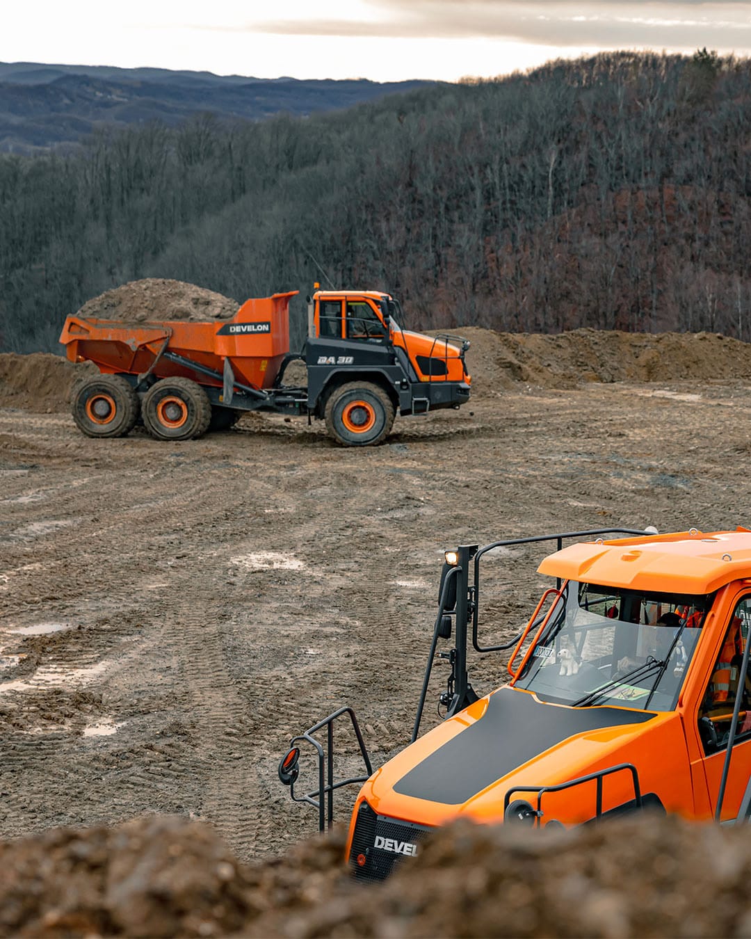 Two Develon DA30 Articulated Dump Trucks operating on a muddy worksite, one in the foreground and the other loaded with material in the distance, set against a backdrop of rugged hills and forest.