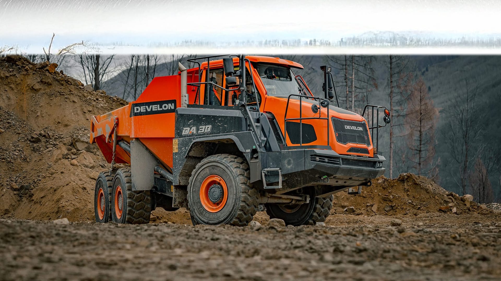 A Develon DA30 Articulated Dump Truck parked on a rugged job site, surrounded by piles of dirt and a backdrop of leafless trees and hills.