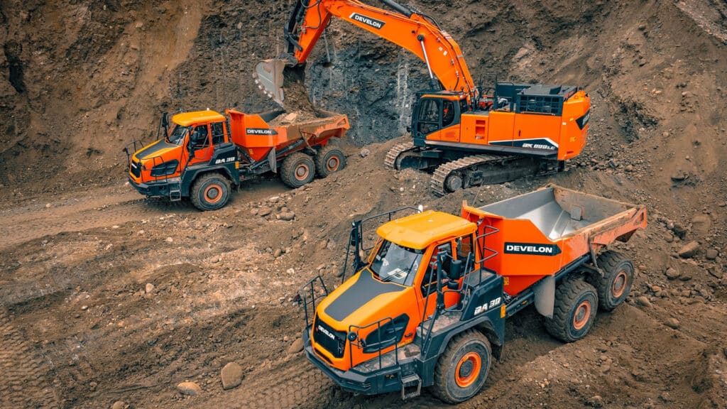 Two Develon DA30 Articulated Dump Trucks working on a construction site, with one being loaded by a Develon DX800LC excavator against a rugged dirt backdrop.